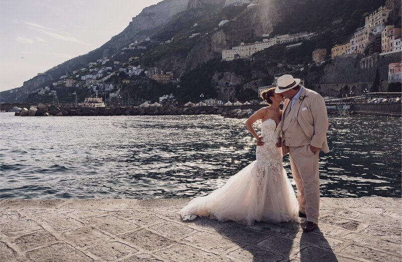 Bride and groom embracing on a seaside stone walkway at a romantic Italy wedding venue, with hills and historic buildings in the background.