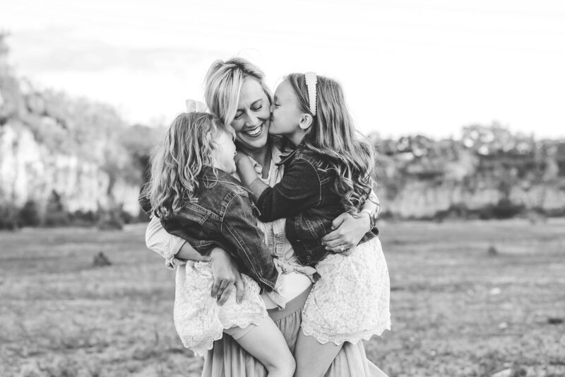 Mother holding both children on each hip while they kiss her cheek