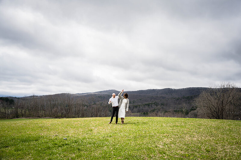A wedding couple holds hands and raises their arms above their head as they bump hips and laugh atop a hill at Heritage Park in Blacksburg, Virginia.