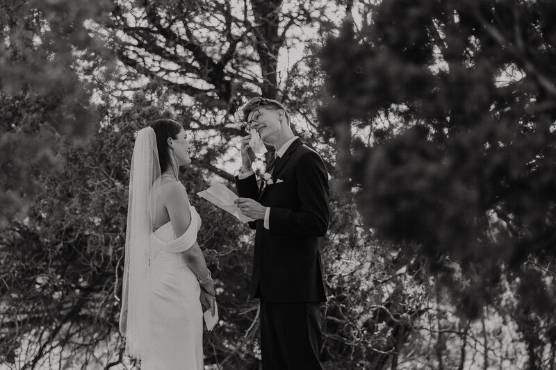 a groom is wiping tears from his cheeks as he reads his vows to his bride standing under pine trees in flagstaff arizona