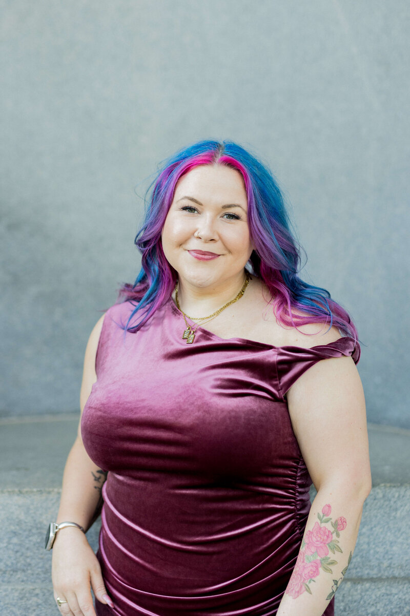 Confident woman with vibrant pink and blue hair poses in a sunlit outdoor setting, wearing a pastel floral embroidered dress with sheer sleeves. Her gold jewelry and bold makeup complement her expressive, creative style. Small business branding photography in Columbia, SC, showcasing unique personal branding for artists, entrepreneurs, and creatives.