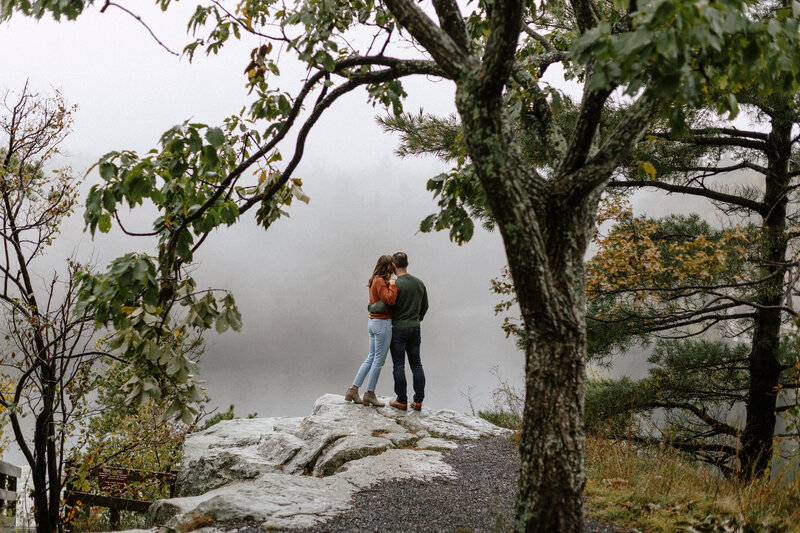 A couple stands on a rocky ledge by a foggy lake, surrounded by trees, sharing an intimate moment in a serene outdoor setting.