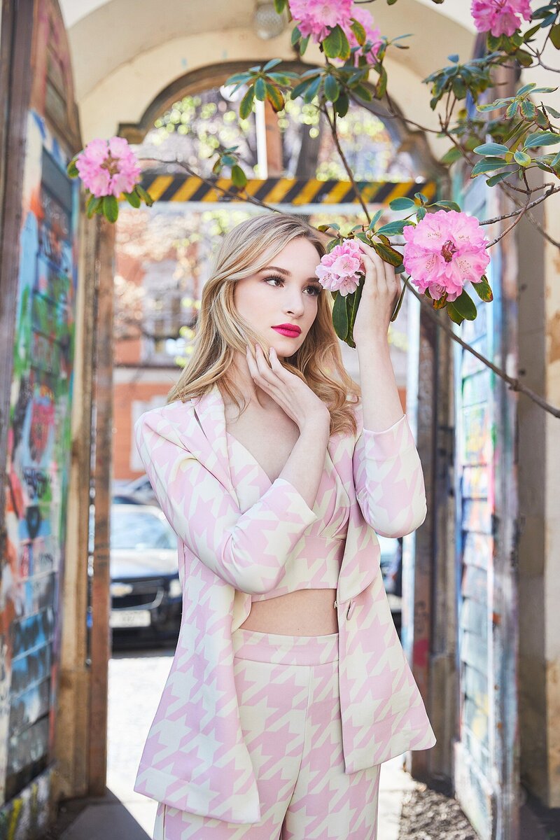 Blonde teen girl in a pink and white houndstooth suit gently holding a pink flower, posing in a vibrant, graffiti-lined urban alleyway in Prague.