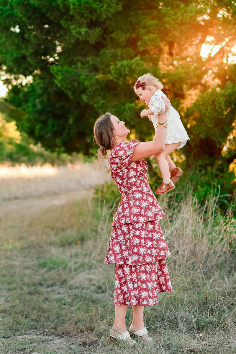 Mother lifting her baby girl during a golden hour family session in Allen, Texas, captured by Jennifer L. Kirk Photography. A joyful motherhood portrait surrounded by nature and warm evening light.