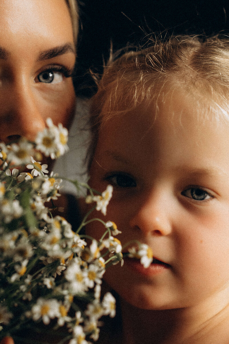 Close-up of a mother and young child surrounded by wildflowers, both gazing softly at the camera in warm, golden light.