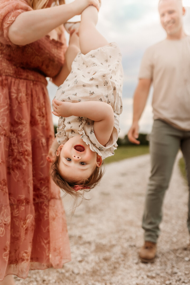 Little girl smiles while mom in pink dress hold her upside down on gravel walkway during a Kansas City Photoshoot.