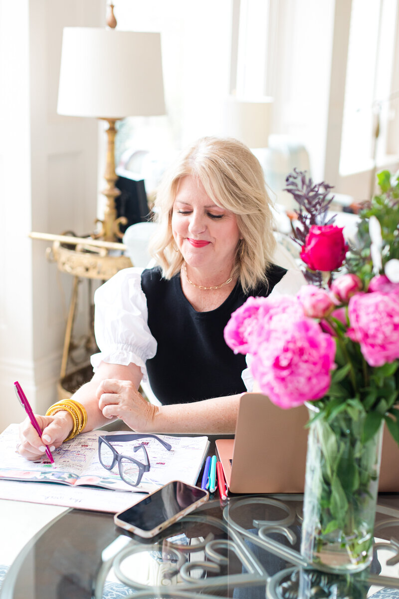Brand photo image of a woman with blonde hair. she is writing down with pink pen and white notebook. on the table are red and pink roses and gold laptop computer. she is wearing a black and white t-shirt