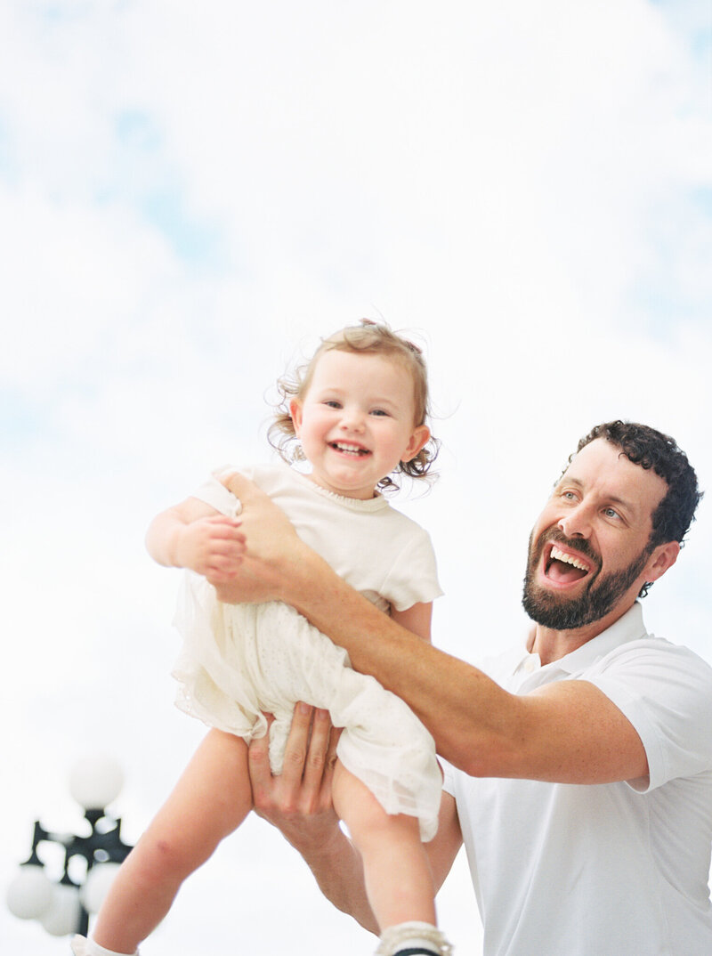 Brunette dad flies young daughter through the air while giggling.