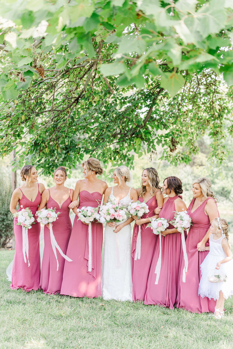 A groom in a pink suit and a bride in her white dress stand hugging each other looking at each other under green trees in a park. Taken by an ohio wedding photographer.