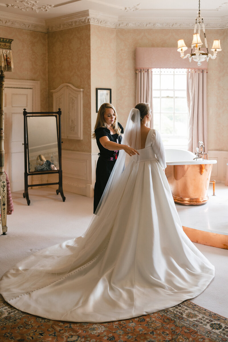 A bride in a white gown with a long train is standing in an elegant room. An attendant helps adjust her dress. The room features a vintage mirror, ornate wallpaper, and a copper bathtub near a window with light pink curtains.