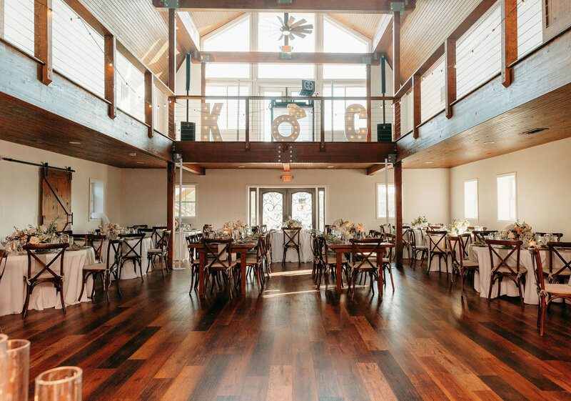 A spacious, sunlit event hall with wooden floors and ceiling, perfect for an elegant reception venue gallery. Tables and chairs are arranged neatly with floral centerpieces. Large windows allow natural light to pour in, and a loft area with balcony railing overlooks the room. A door is centered at the back.
