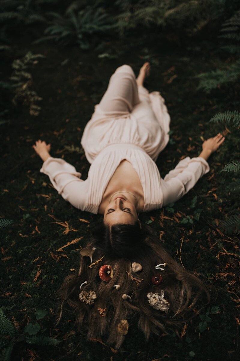Woman with long, brown hair lays on forest floor with her hands lay by her side. Small mushrooms and foliage are laid flat across her outspread hair.