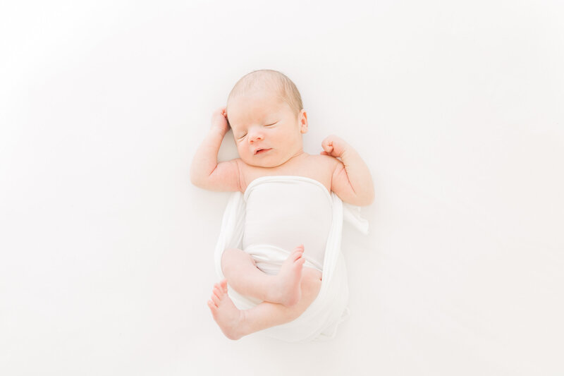 A newborn baby sleeps in a swaddle on a white bed