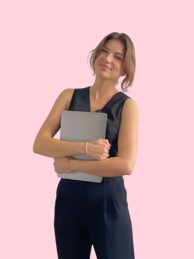 Caitlin Christensen holding a laptop with a relaxed smile. She wears a black vest and black trousers against a pink background.