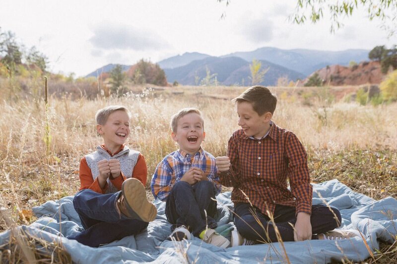 Three boys are laughing seated in the yellow grass with the front range in the background.
