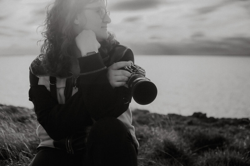 The photographer is crouched on a grassy hill with a lake behind her. She's holding a camera and looking to the right.
