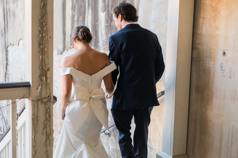 A bride in an off-the-shoulder white gown with a large bow walks down stairs holding hands with a groom in a dark suit, both facing away from the camera beside a textured, weathered wall.
