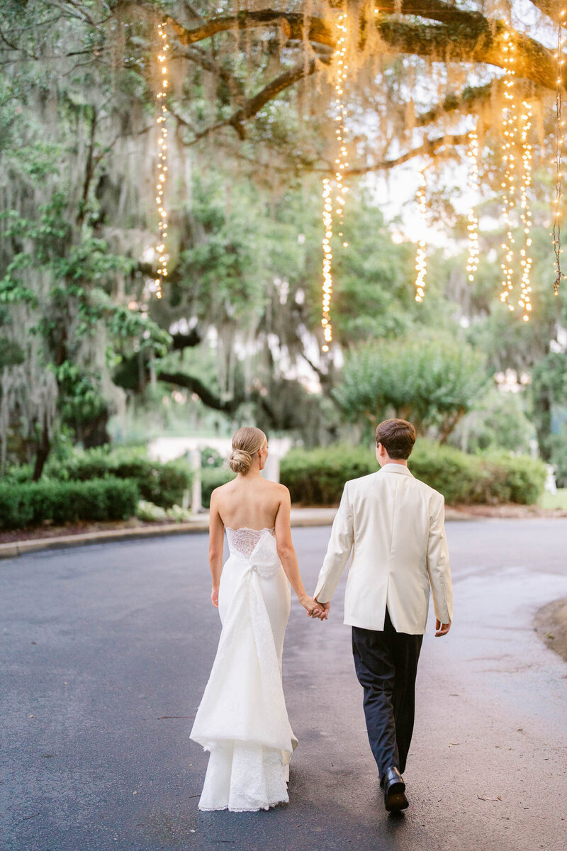 Bride and groom walking hand in hand under hanging string lights surrounded by trees at their Myrtle Beach wedding