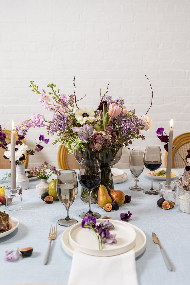 Beautiful table setting for a wedding featuring a white table cloth, dark wine glasses, purple, green and white floral bouquets, and figs