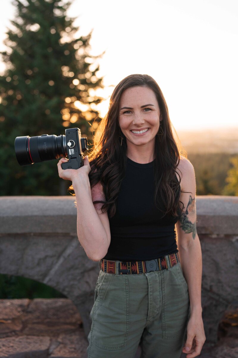 a female photographer smiles at the camera while holding a camera in her hands during golden hour