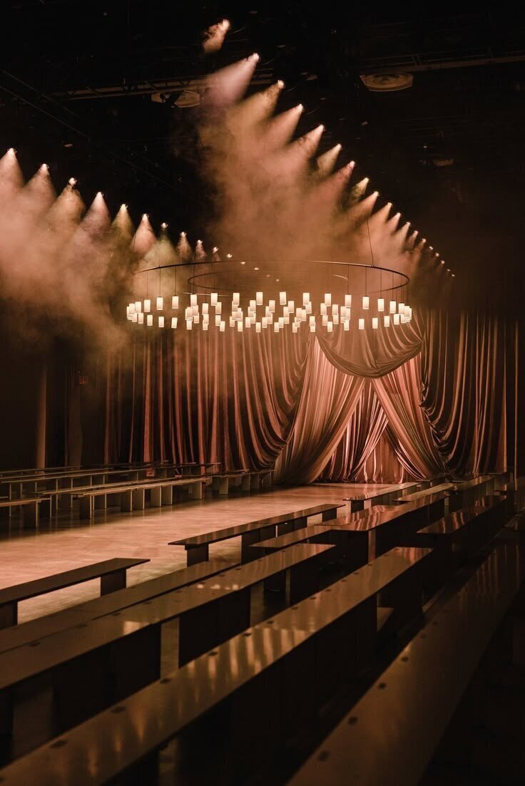 Dimly lit elegant interior with benches, a circular chandelier, and flowing drapery.