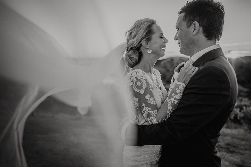 A black and white image of a bride and groom being windswept with the veil blowing in the wind during a wedding day photoshoot with Tashina Narelle, Auckland and Waikato Wedding Photographer