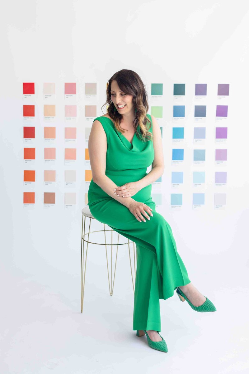 Color psychology expert in emerald green romper sharing authentic smile while seated on gold stool against wall of organized color swatches