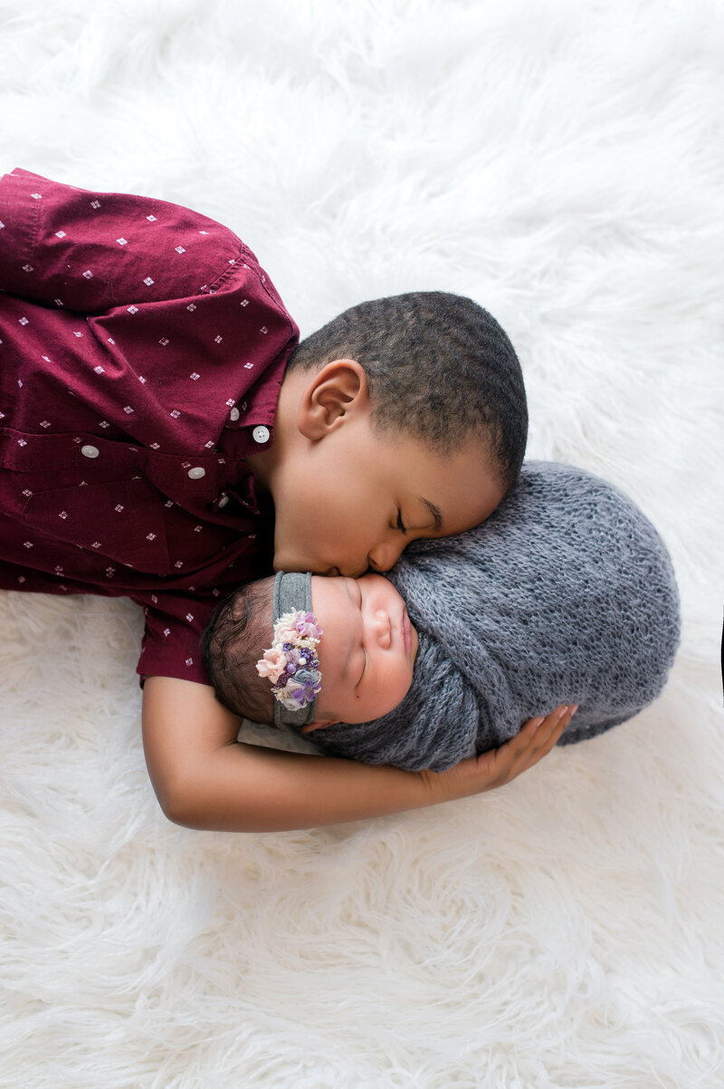 Toddler boy kissing his newborn baby sister during posed studio portrait session.
