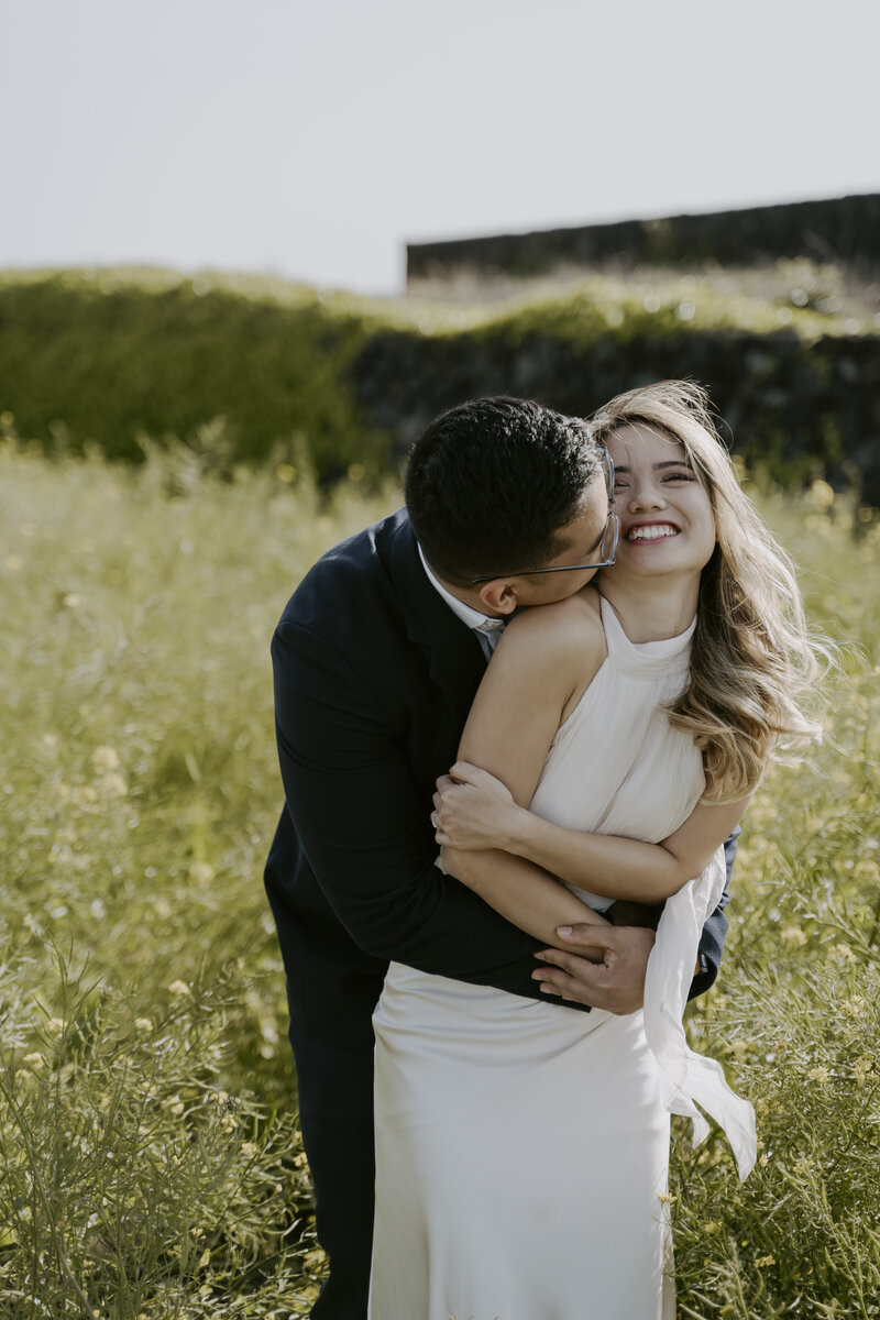 a man in a blue suit hugs a woman in an ivory flowy dress from behind during their prewedding shoot