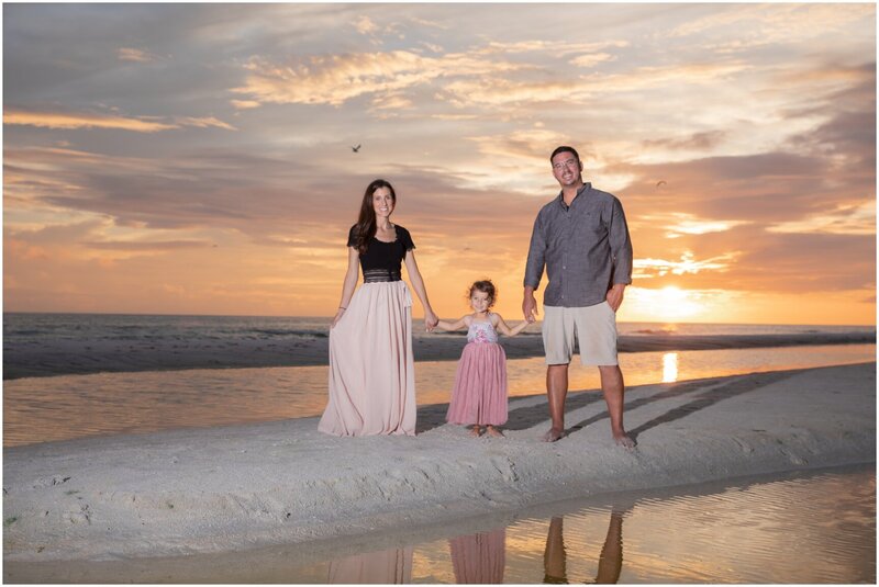 A couple and their young daughter holding hands on the beach in front of a gorgeous sunset.