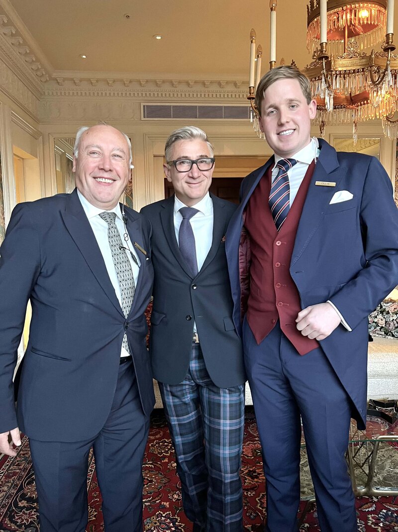 Three well dressed men standing together with a fancy chandelier behind them 