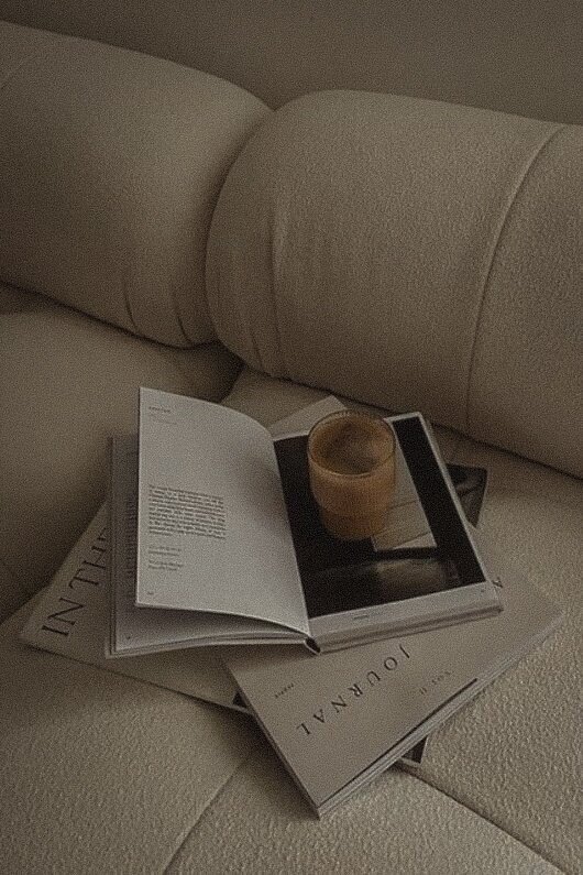 Neutral-toned flatlay of an open journal, coffee in a glass cup, and books on a textured lounge, reflecting the calm, intentional aesthetic of Aurum Design Studio.
