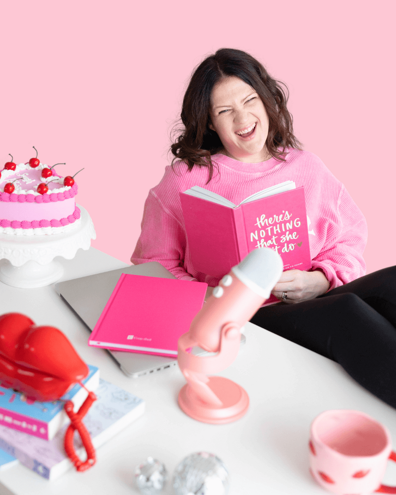 Emily, a brand and web designer for women, sits at a pink table, wearing a cozy pink sweater holding a journal that says "there's nothing she can't do."