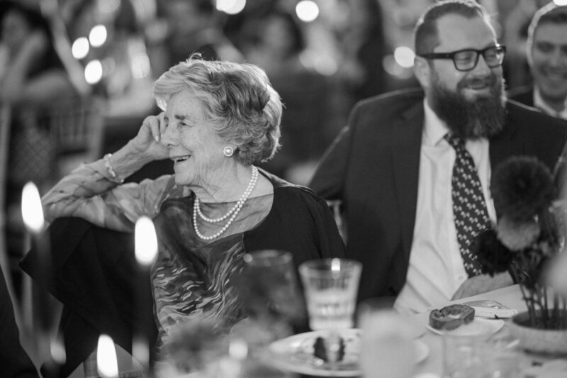An elegantly dressed grandmother wearing pearls sits at a table with blurred guests, illuminated by candles and festive decorations.