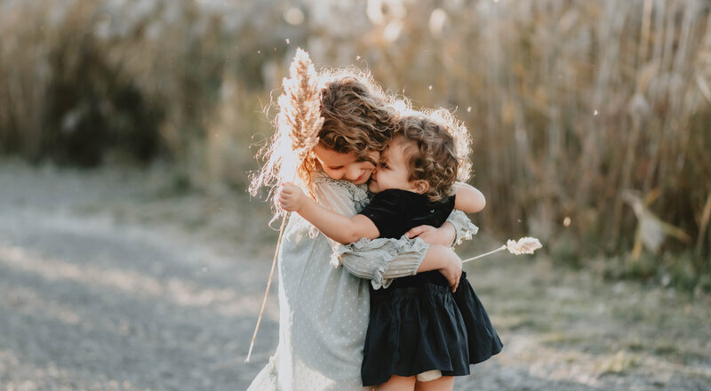 sisters hugging in golden hour and laughing during family photos in a windsor ontario field