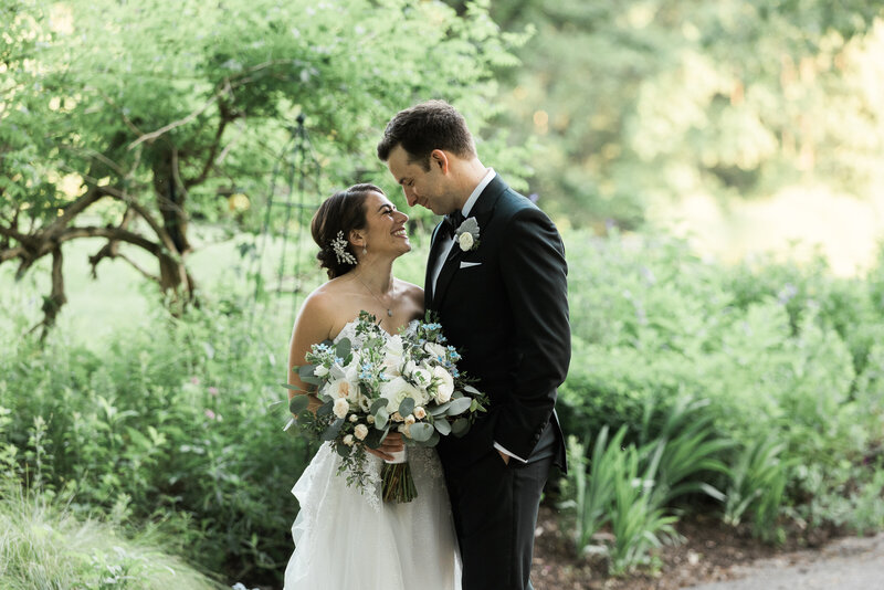 a white groom in a black tux facing and holding a white brunette bride in a strapless white gown holding her white and blush bouquet smiling at one another