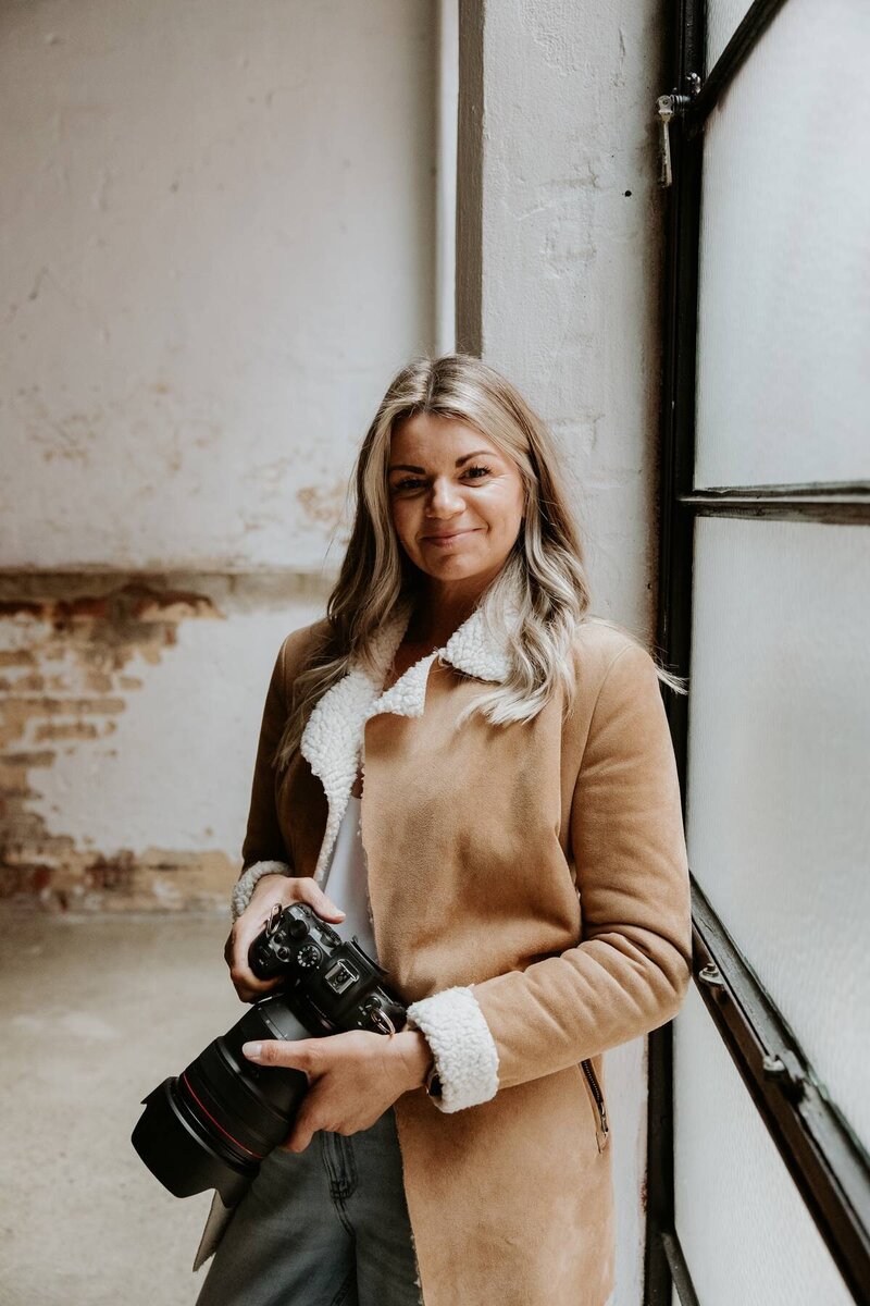 Photo of Kaz Soom, Perth photographer, smiling at the camera whilst holding her own camera.  She is wearing a shearling jacket. 