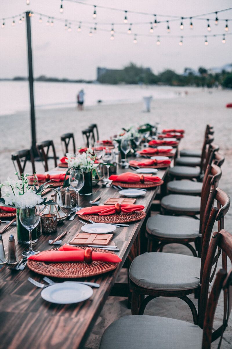 Chairs and a rectangular dining table designed with red napkin, utensils and glasses beside the seashore