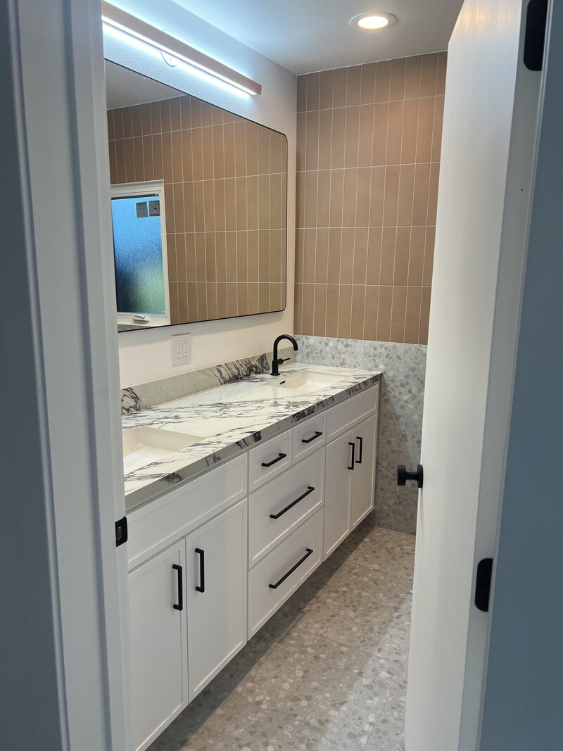 A remodeled bathroom with terra cotta subway tile on one wall, a white vanity with marble looking tops and a large mirror.