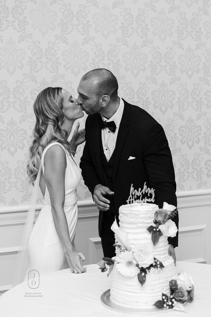 Black and white image of a couple kissing behind their wedding cake.