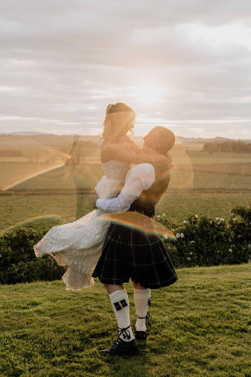 A groom lifts a bride in the air and he twirls her around in circles. The golden evening sun shines and rain falls all around. The photographer's lens is covered in water and the wedding photo takes on a dreamy quality.
