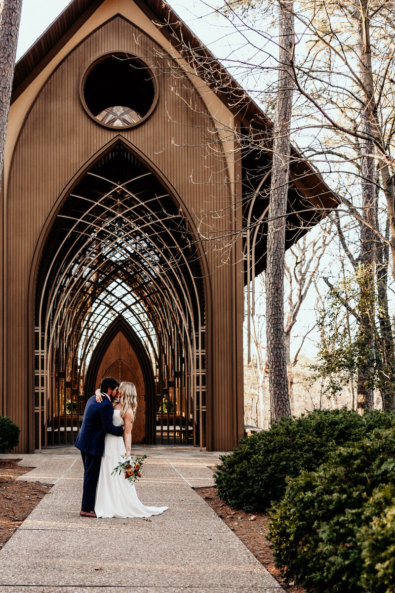 The bride and groom embracing and kissing each other in front of the glass chapel in Bella Vista, AR