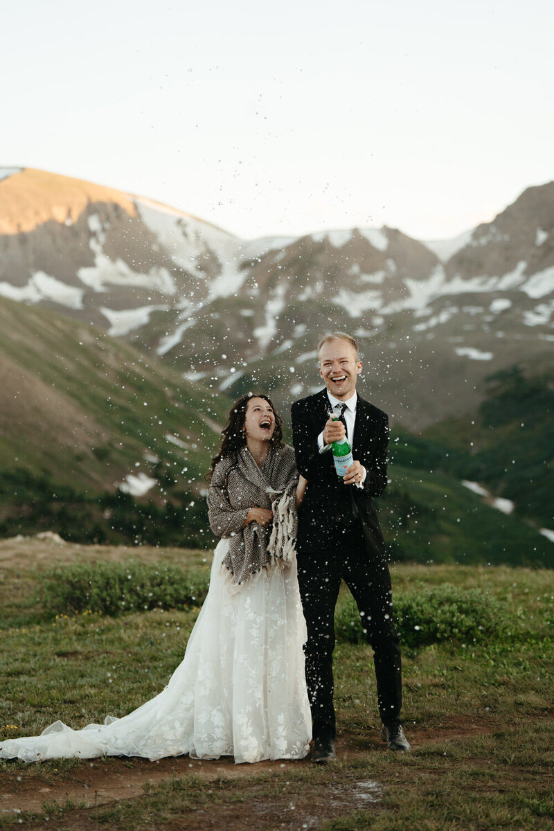 Bride and groom pop champagne on a mountain during their hiking wedding with big smiles on their face