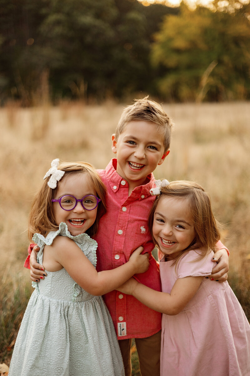 Three young siblings sharing a joyful hug during a golden-hour family photography session in West Michigan, capturing genuine smiles and warm connections