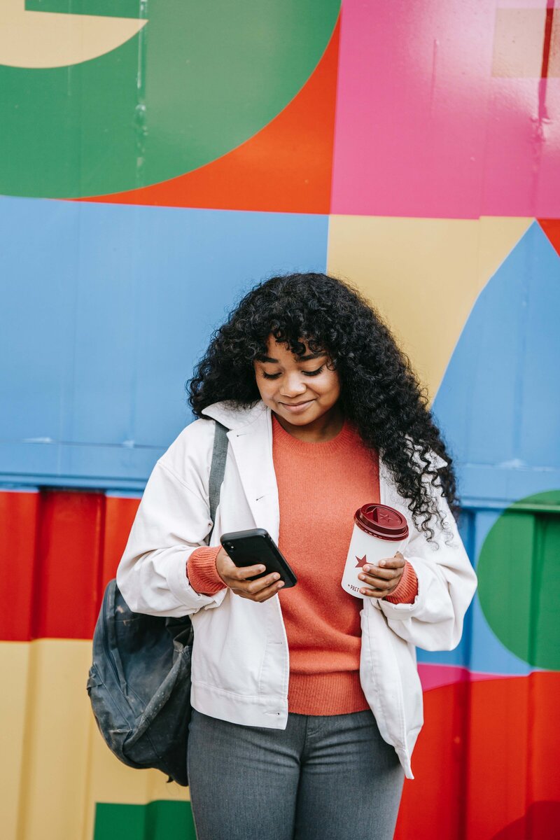 This image shows a young feminine-presenting person of color standing outside, near a colorful mural. They are holding a coffee cup in one hand and their phone in the other, smiling down at it.