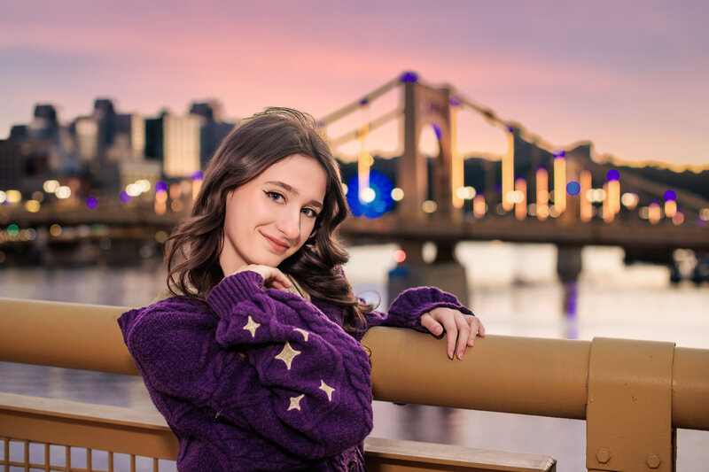 Identity senior girl wearing a bright purple sweater is offset by a fluorescent pink Sunset sky over the bridges in Pittsburgh Pennsylvania.