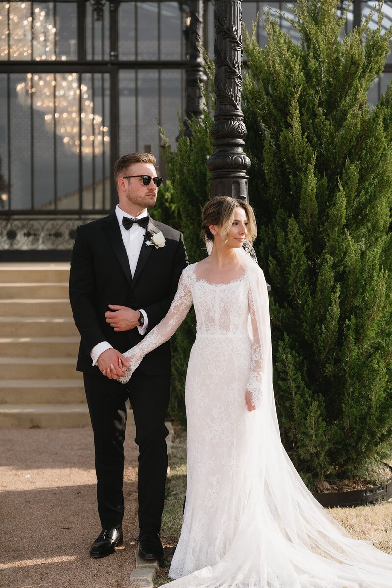Bride and groom holding hands outdoors, bride in lace gown and groom in tuxedo.