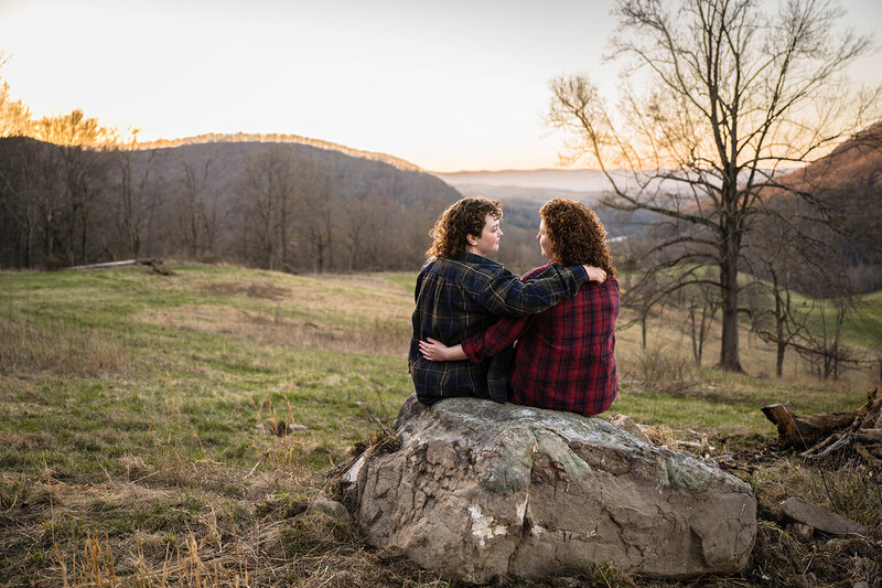 A queer couple sit on a large rock and look at one another as the sun begins to set behind the Blue Ridge Mountains.