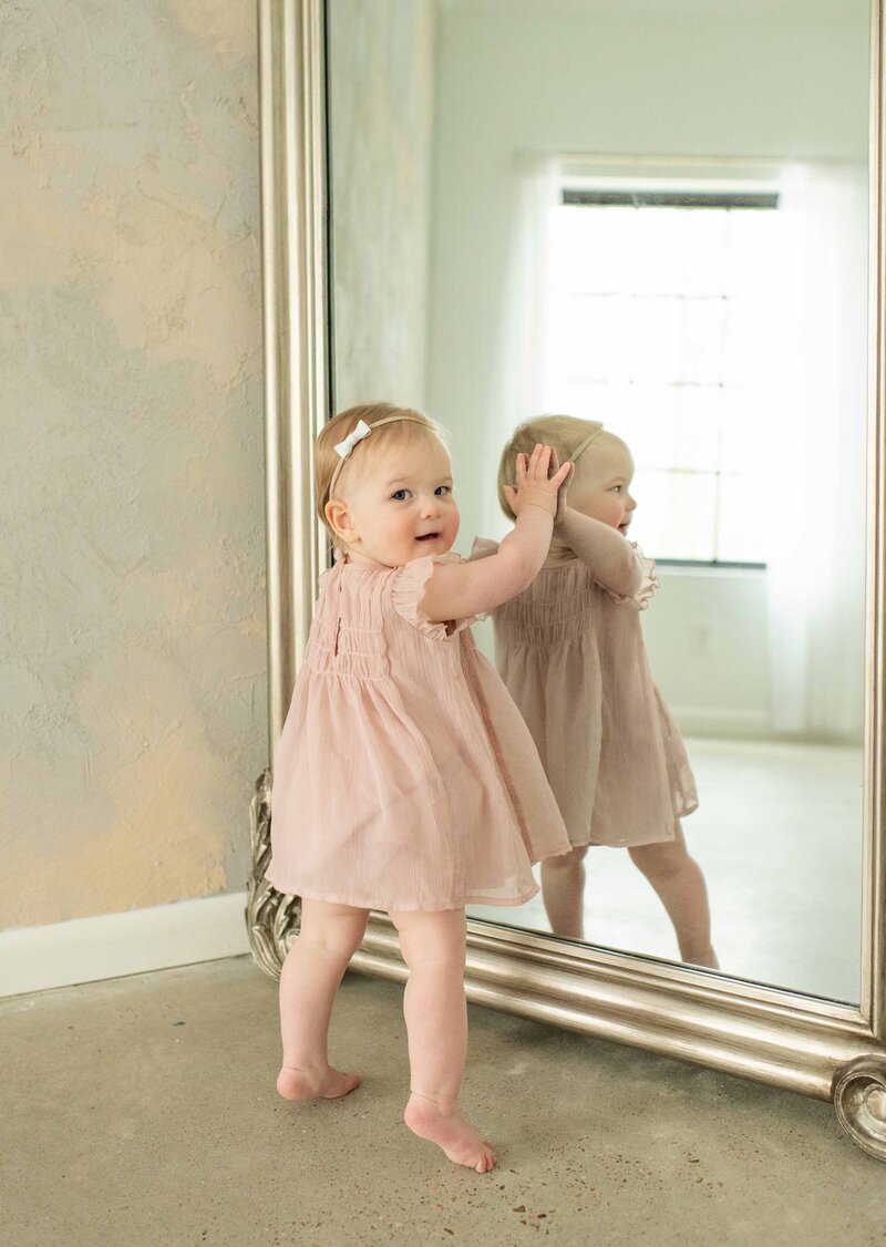 Toddler in a pink dress reaches for her reflection in a mirror, smiling. A playful baby session by Moments in Light Photography.