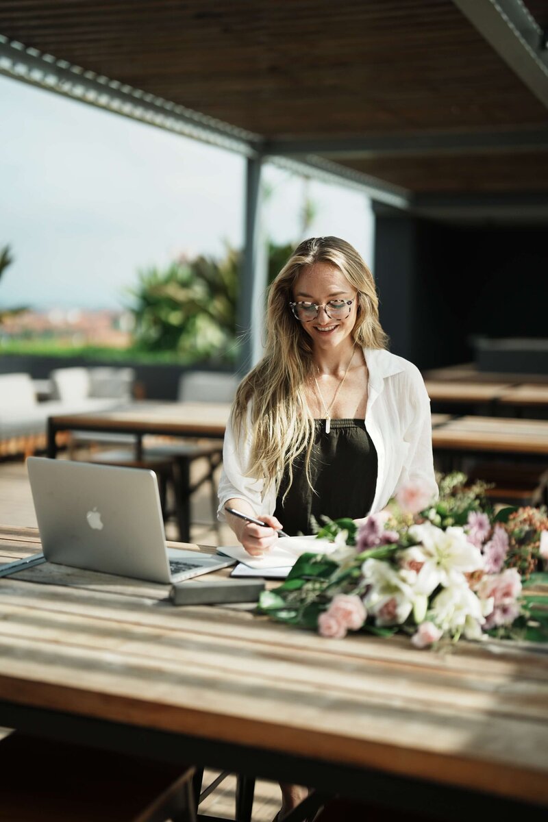 Hypnotherapist, RTT® Practitioner, RMT and RYT Noelle Rivet writing  in a journal at a desk with her laptop